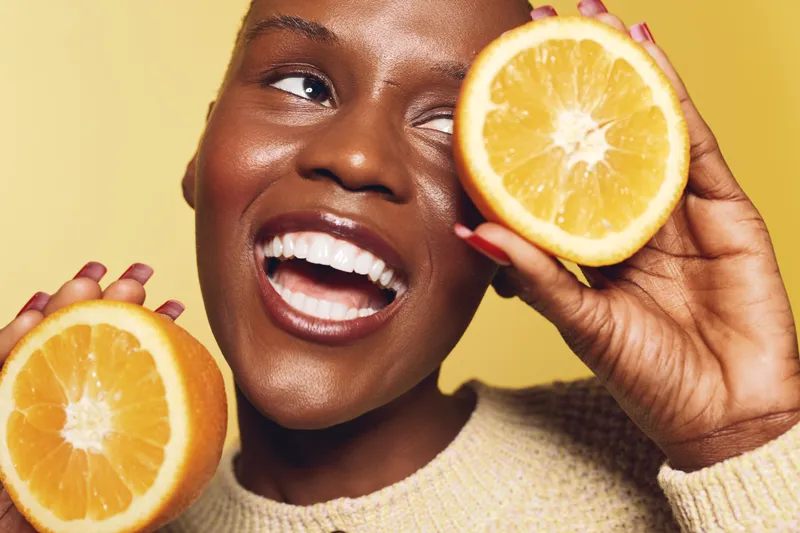 Happy dark skinned African woman smiling broadly holding two halves of fresh orange close to her face. Concept of healthy lifestyle, vitamin C, wellness, and natural beauty with citrus fruit.