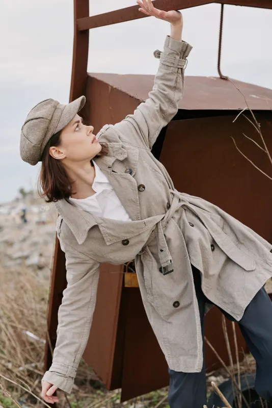 Young woman in beige trench coat and cap poses outdoors near rusty metal structure, looking up with arm raised. Fashion concept with natural light and muted autumn colors.