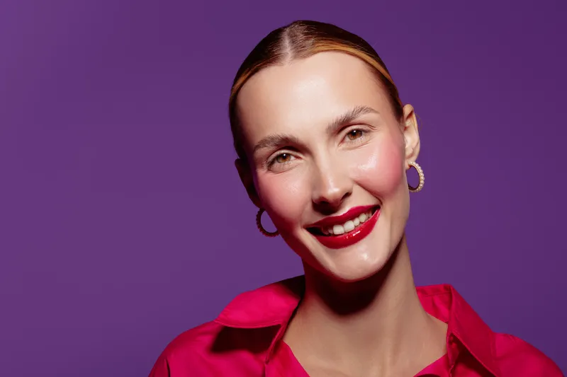 Smiling woman with red lipstick and hoop earrings wears bright pink shirt against purple background. Close-up portrait with natural skin glow and confident expression. Concept of beauty and fashion.