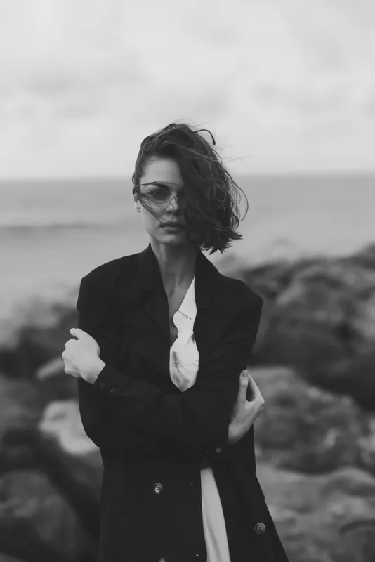 Woman in fashion jacket by the ocean, wind tousling hair, monochrome mood and moody outdoor scene, showcasing stylish attire against a windy coastline