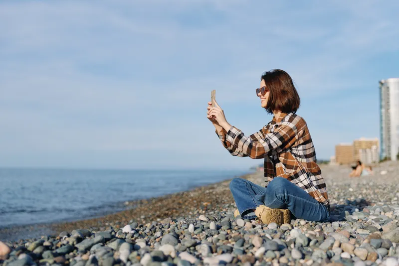 Woman on a pebble beach takes a selfie with her smartphone, wearing a plaid shirt and jeans, as waves meet the shore and a distant cityscape creates a calm coastal scene.