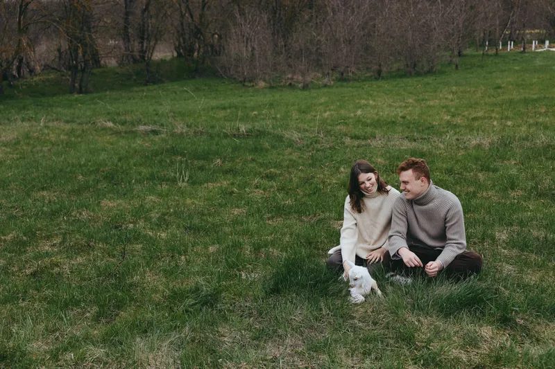 Young couple sitting on green grass in casual sweaters enjoys spending time with a playful baby goat in a peaceful outdoor setting during early spring.