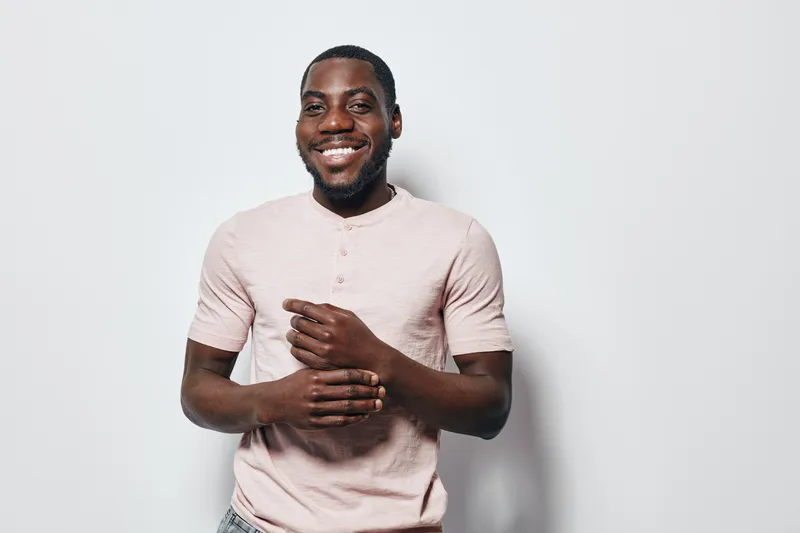 African American man with dark skin smiling and standing against white background wearing light pink shirt. Portrait of happy male with beard and casual style in studio.