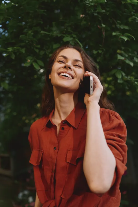 A smiling woman on a mobile call in an outdoor setting, wearing a rust shirt. Lush greenery in the background highlights a casual lifestyle moment and everyday communication