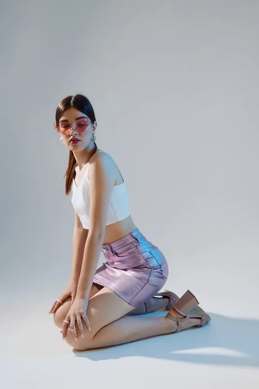 Young woman in white top and shiny purple skirt kneels on the floor posing. She wears sunglasses and heels in a studio with soft lighting. Concept for beauty fashion style.