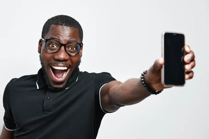 African American man with dark skin and glasses smiling excitedly while holding smartphone for selfie or video call in studio with white background and casual black shirt.