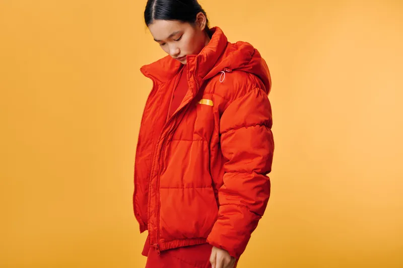 Young woman wearing orange puffer jacket standing against yellow background looking down. Fashion, winter clothing, casual style and warm outerwear for cold weather season outfit.