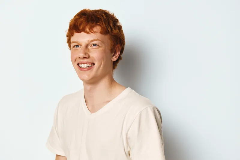Young redhead man smiling and looking away in white t shirt on plain light background for casual portrait with natural light and happy mood.