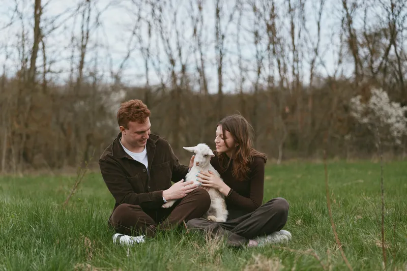 Happy couple in a grassy field with a playful goat kid shares a tender moment outdoors, showcasing simple rural joy and friendly animal interaction in natural light on a mild afternoon.