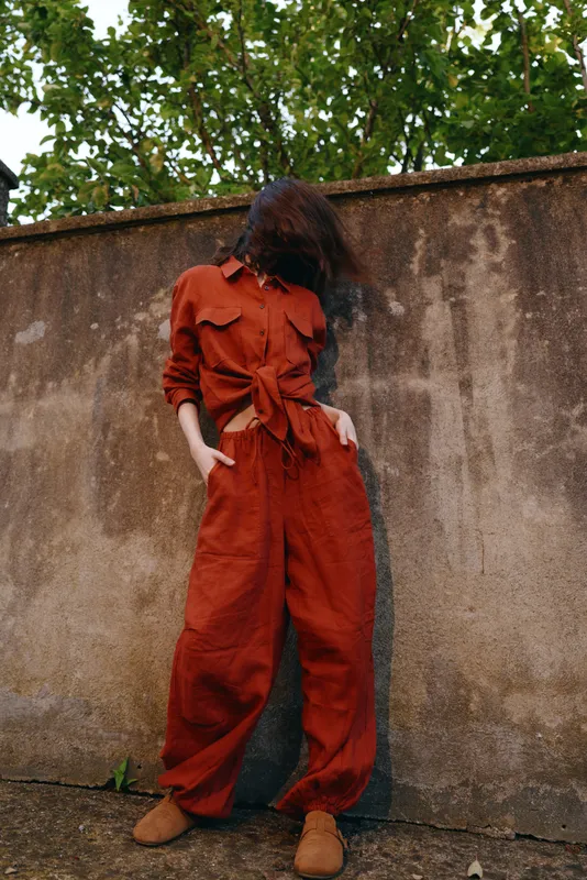 Young woman wearing a rust red jumpsuit stands against a weathered wall, wind tousling hair, hands in pockets, exuding moody urban confidence