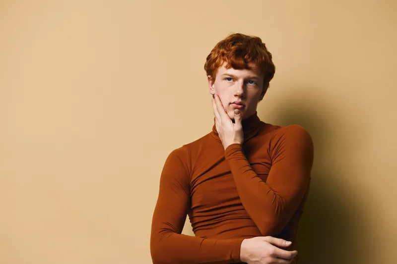 Young man with red hair wearing brown turtleneck posing thoughtful on beige background. Portrait of male model with hand touching face in studio with soft light and shadow.