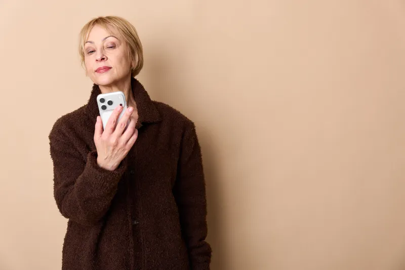 Elderly woman holding smartphone indoors portrait, wearing dark fuzzy coat against beige wall, relaxed expression for communication, while texting, soft studio lighting, minimal background, copy space