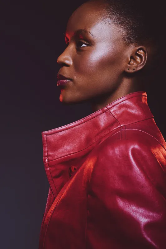 Profile of a confident black woman with dark skin wearing a red leather jacket. Studio portrait with dramatic lighting on a dark background showcasing fashion and beauty concepts.