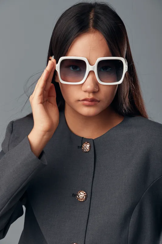 Young woman in oversized white sunglasses and gray blazer poses confidently with serious expression. Portrait fashion style with long dark hair on neutral background in studio lighting.