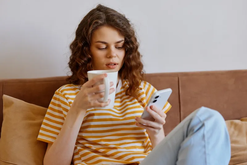 Young woman sits comfortably on a soft sofa indoors, sipping a warm mug and scrolling her phone, enjoying a calm, reflective moment.