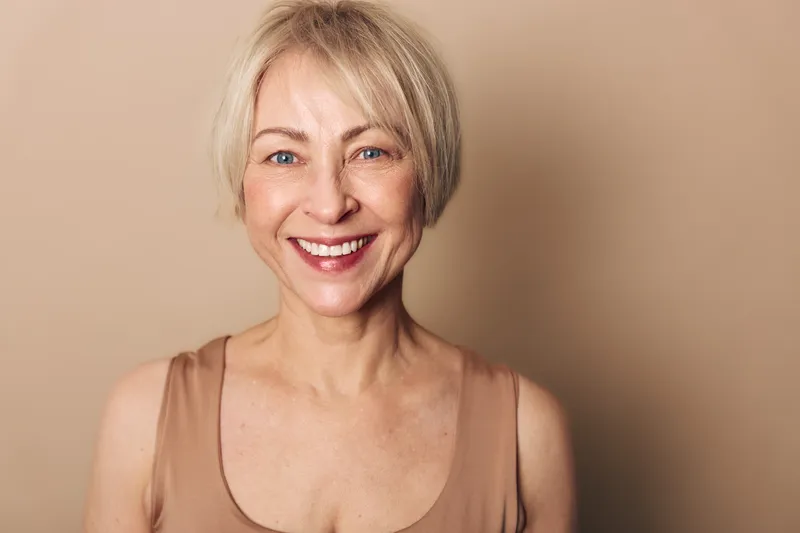 Smiling middle aged woman with short blonde hair and natural makeup wears beige top on beige background. Concept of happiness, aging beauty, confidence, and positive lifestyle.