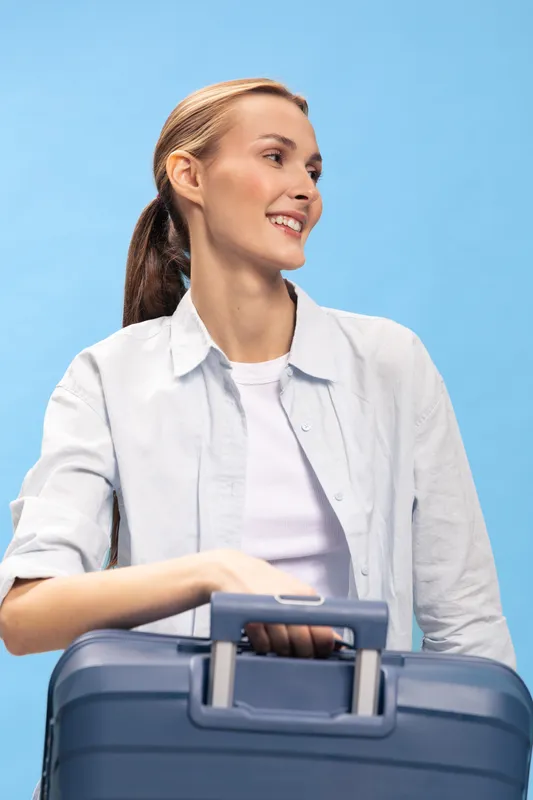 Young woman with ponytail holding blue suitcase smiles looking to side on light blue background. Concept of travel, vacation, and positive emotions.