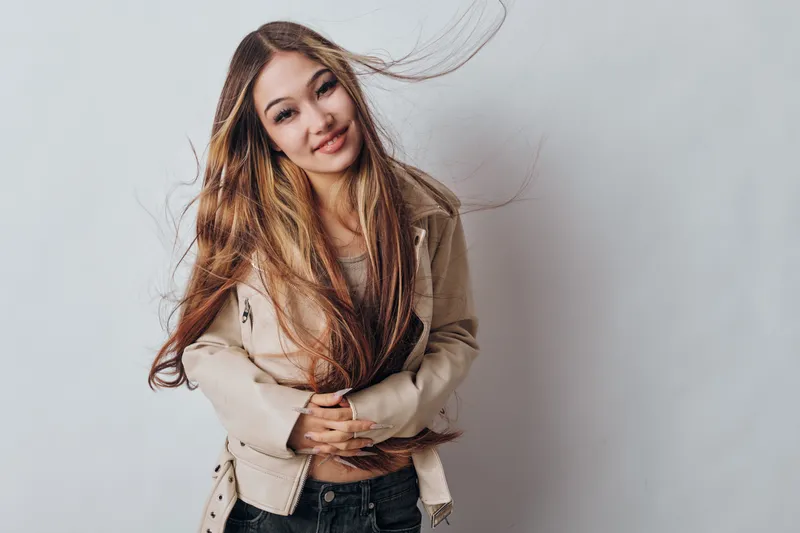 Young woman with long hair smiling and posing in casual beige jacket and jeans with flying hair on light background indoors. Female model, fashion and beauty with natural look and confidence.