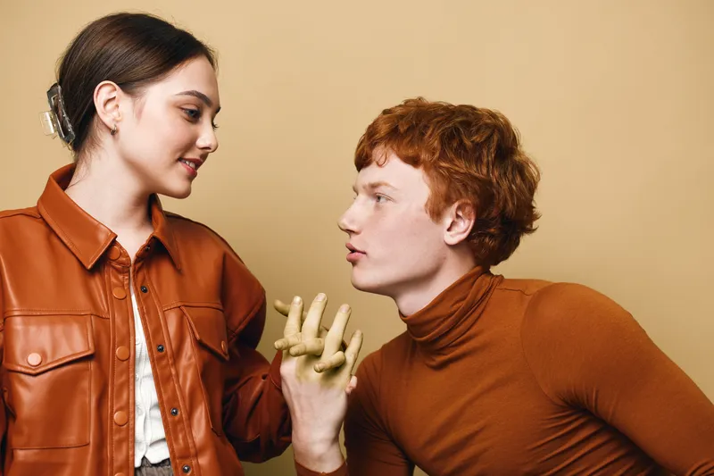 Young woman and man holding hands with golden paint on fingers, looking at each other with smiles and connection in stylish casual clothes on beige background.