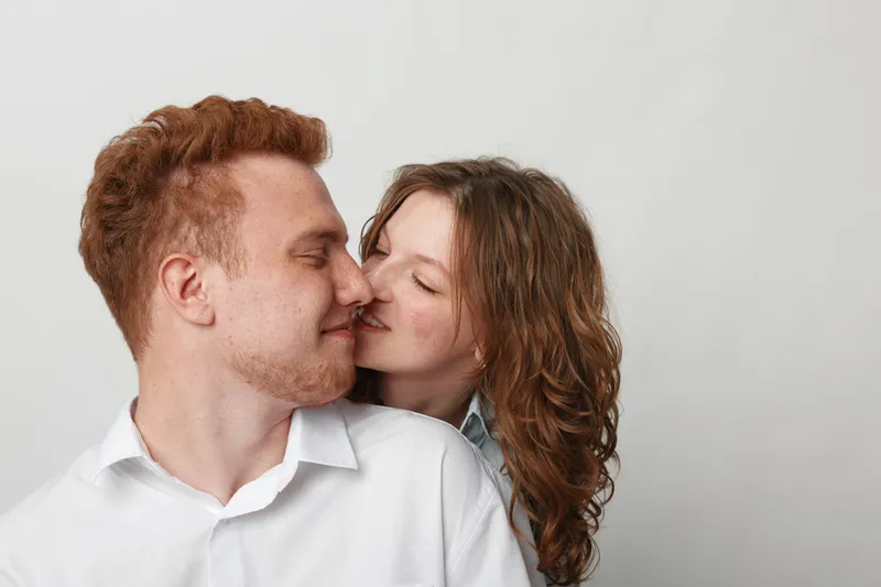 Young couple in white shirts shares a tender moment with closed eyes and gentle smiles against a plain light background. Concept of love, intimacy, and affection.