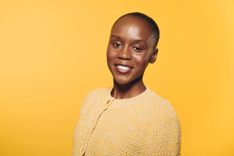 Smiling young black woman with dark skin wearing yellow sweater stands against bright yellow background. Portrait of confident African female model with natural makeup and short hair.