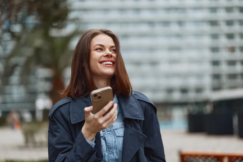 Young woman outdoors, smiling while checking her phone, wearing a denim dress and navy jacket in a casual urban park scene, soft light background urban.