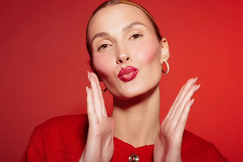 Young woman with red lipstick and red sweater blows a kiss on red background. Close-up portrait with hoop earrings and natural makeup. Concept of beauty, affection, and fashion.