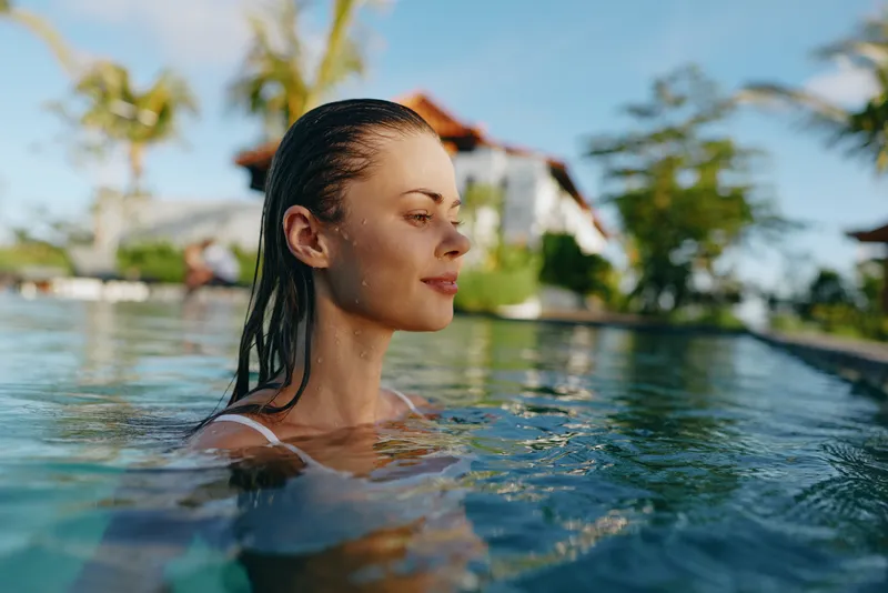 Relaxed woman in a pool, sunlit water glistening, tropical backdrop, showcasing lifestyle, vacation, wellness, and self care in an outdoor pool scene with soft light and warm mood.