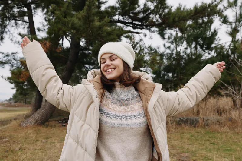 Joyful woman in a beige puffy coat and beanie stands outdoors with arms raised, smiling in a park setting. Cozy sweater visible, autumn trees and open space in the background.