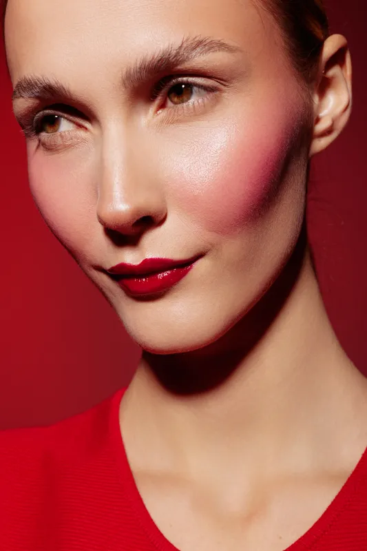 Close-up portrait of a woman with red lipstick and pink blush on a red background. The image highlights makeup, beauty, and fashion concepts with soft lighting and smooth skin texture.