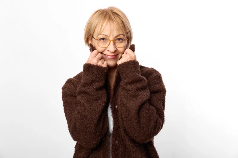 Elderly woman with glasses smiles, covering mouth with hands in a brown coat, studio portrait on white background, calm friendly gesture, winter fashion.