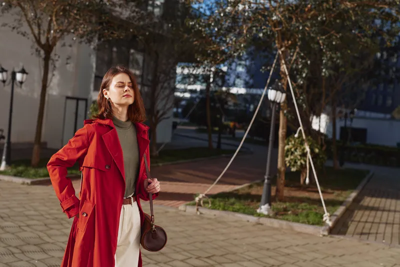 Young woman in a red coat stands confidently outdoors with closed eyes enjoying the sunlight on a clear day in an urban setting.