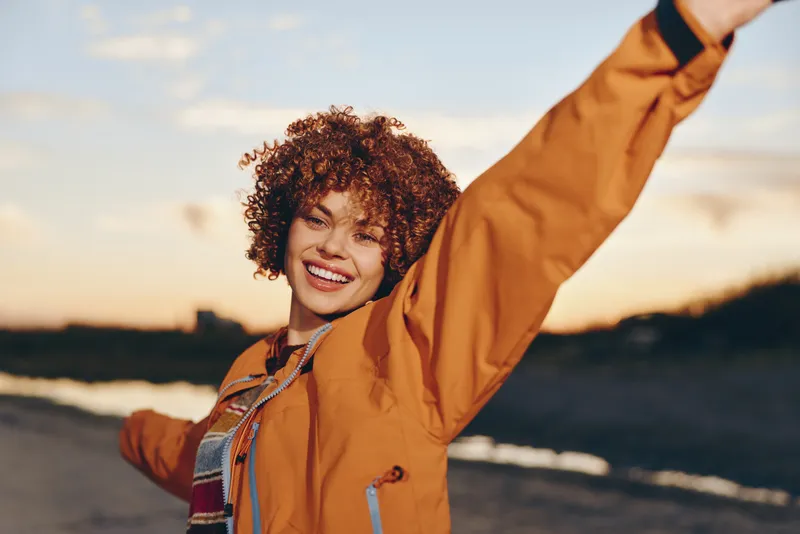 woman lifestyle outdoors smiling curly hair in orange jacket at sunset by riverbank, one arm raised, casual travel, warm golden light, soft breeze, scenic nature calm background