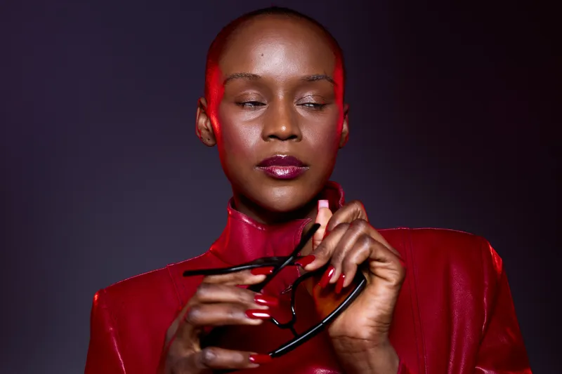 Confident African woman with dark skin wearing a shiny red jacket holds black sunglasses, posing against a dark background. Concept of fashion, elegance, and modern style.