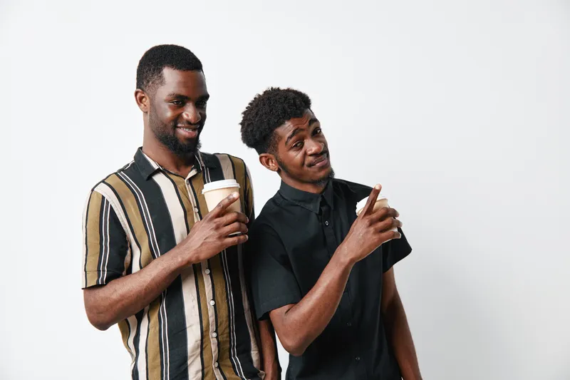 Two young African American men with dark skin smiling and holding coffee cups, standing together on white background in casual clothing for friendship and lifestyle.