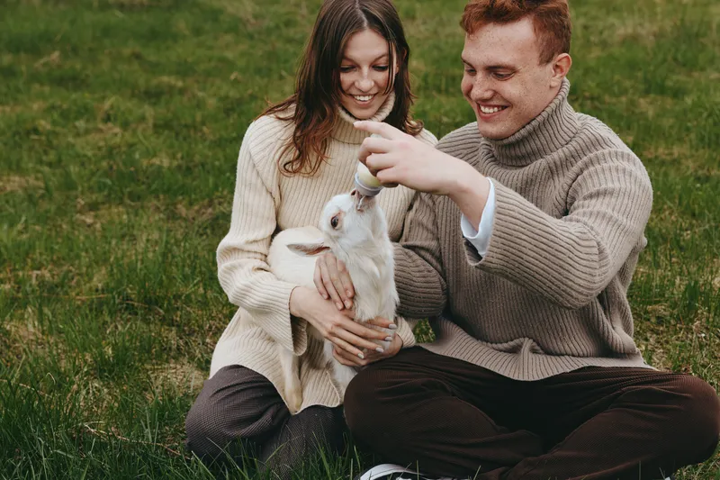 A young couple in cozy sweaters sits on grass while feeding a baby goat with a bottle, enjoying a relaxed moment in nature and bonding with the animal in a casual lifestyle setting.