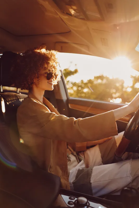 A stylish woman with curly hair drives a car in warm retro tones, sunglasses on, sunlight streaming through the window. A lifestyle portrait capturing carefree travel moments.