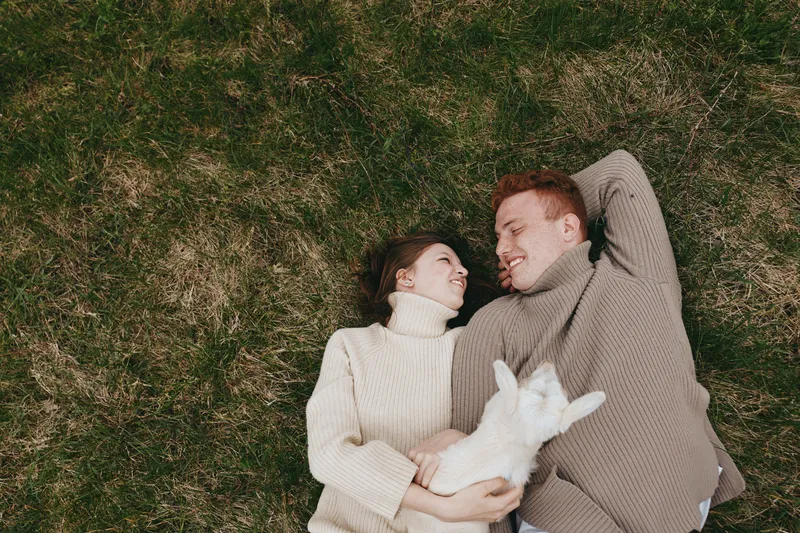A couple lies on grass, smiling, while a playful goat kid rests in their arms. A lifestyle moment highlighting affection, nature, and gentle animal companionship.