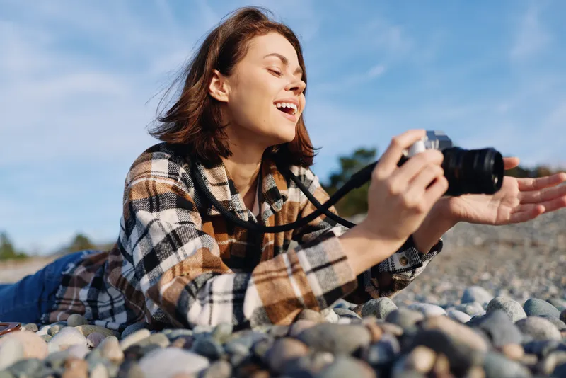 Woman smiling while lying on pebbles at the beach, holding a camera for candid photography outdoors. A relaxed lifestyle moment captured in natural light, showcasing hobbyist creativity and joyful