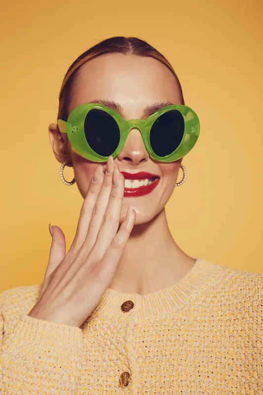 Smiling woman with green round sunglasses and red lipstick covers mouth with hand. She wears yellow knitted sweater and silver hoop earrings on yellow background. Fashion and style.