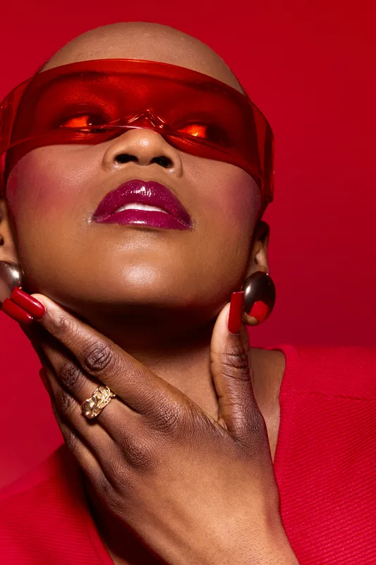 Close-up of a confident black woman with dark skin wearing red translucent glasses and matching lipstick and nails. Fashion concept with bold makeup and accessories on red background.
