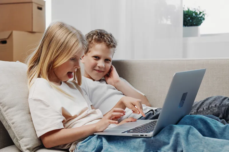 Two children sitting on a couch using a laptop together, one excitedly pointing at the screen. Bright room with natural light, cardboard boxes in the background suggest moving or unpacking.