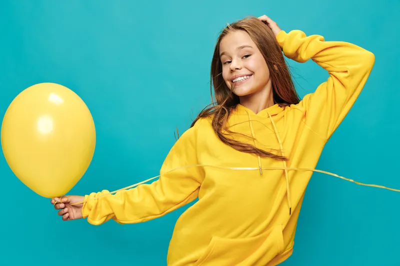 Girl in yellow hoodie smiling and holding yellow balloon on blue background. Happy teenage female with long hair posing and enjoying colorful casual style and cheerful mood.