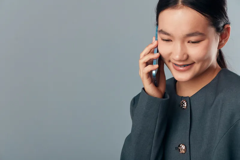 Young woman with dark hair talking on mobile phone against grey background, smiling and wearing dark grey jacket with decorative buttons in closeup portrait photo.
