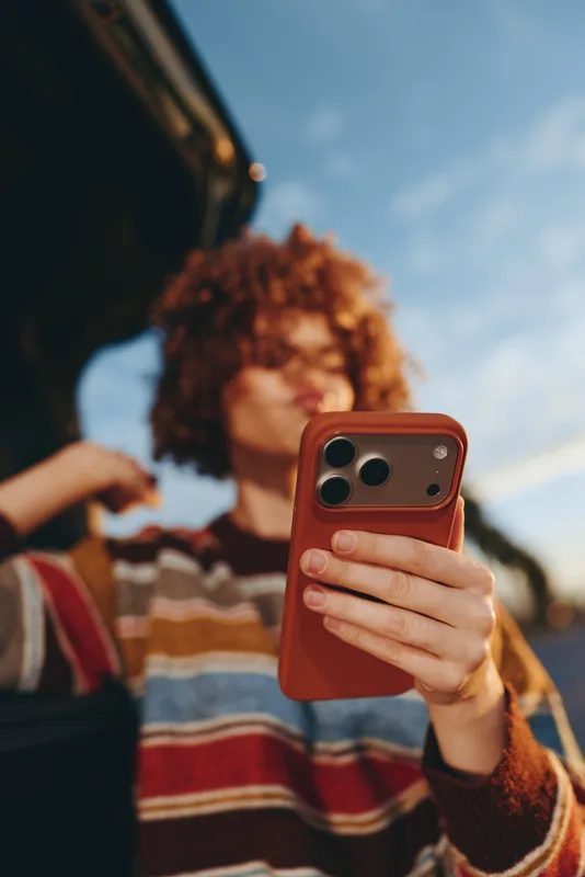 Woman lifestyle outdoors, holding smartphone for selfie, curly hair visible, casual striped sweater, sky background, soft daylight blur, relaxed street scene, warm smile