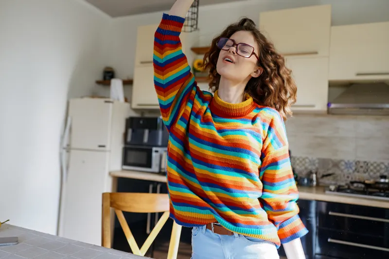 Cheerful woman indoors wearing a colorful striped sweater, enjoying a casual home moment in the kitchen, relaxed pose, natural light, vibrant colors, lifestyle vibe