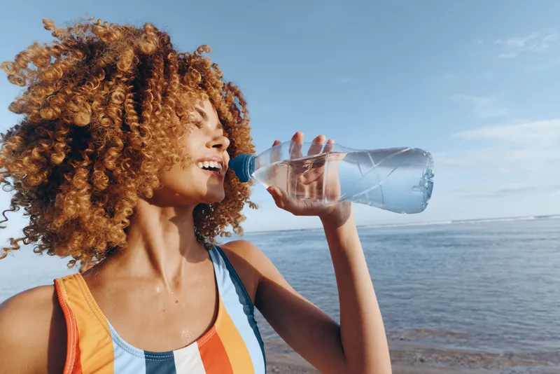 Curly woman drinks water from plastic bottle near sea on sunny day, wears colorful striped top, enjoys fresh hydration and outdoor summer lifestyle, positive mood