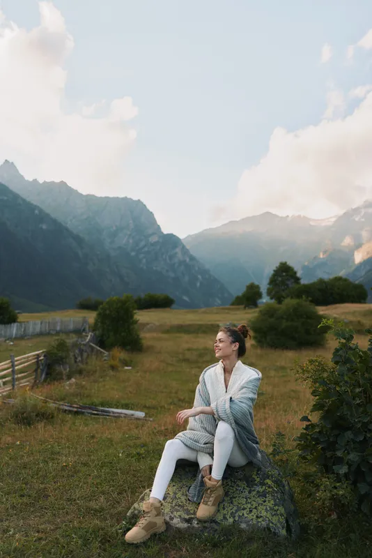 woman enjoying nature in mountainous meadow, sits on rock, wearing cozy cardigan and boots, peaceful outdoor lifestyle portrait with scenic landscape and natural light