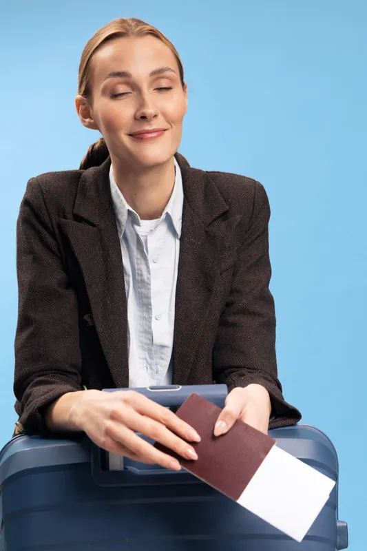 Smiling woman holds passport and boarding pass above suitcase on blue background. Travel concept with happy female traveler preparing for trip, vacation, and holiday planning in studio.