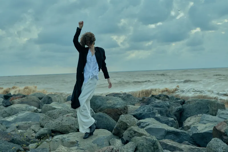 Figure on rugged rocks by the sea, wearing a long dark coat and light trousers, striking a dynamic pose against wind and cloudy skies, capturing movement and resolve